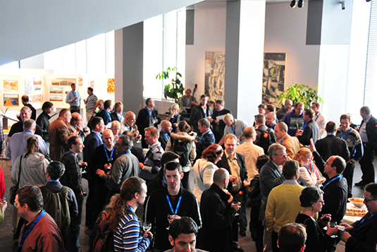 A large group of people are gathered in a conference room. Some individuals are conversing, while others are mingling and holding drinks. The room features artwork on the walls and large windows in the background, providing a bright and airy environment.