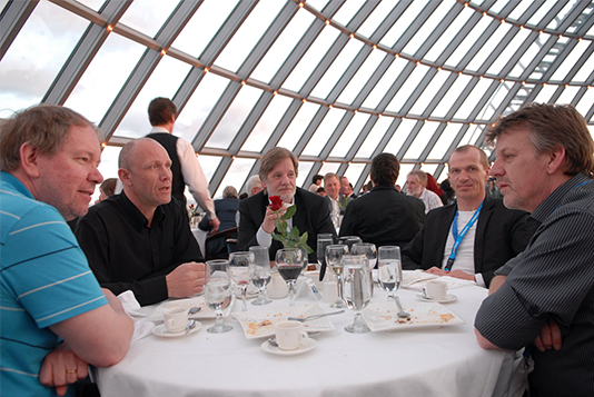 A group of five men sitting around a table, engaging in conversation and collaboration. More people are visible in the background.